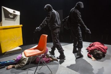 Kaksi mustiin vaatteisiin pukeutunutta naamioitunutta henkilöä, joista toisella on ase kädessään, kävelee kahden maassa makaavan ihmisen ohi, joista toinen on osittain oranssin tuolin alla ja toinen käpertyneenä. Etualalla on suuri keltainen laatikko.