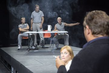 Kolme harmaisiin asuihin pukeutunutta miestä istuu näyttämöllä pöydän ympärillä, kaksi istumassa ja yksi seisomassa, ja ilmassa on savua. Savukkeella oleva mies ja nainen katsovat kohti lavaa etualalla. Lavastus vaikuttaa teatraaliselta ja dramaattiselta.