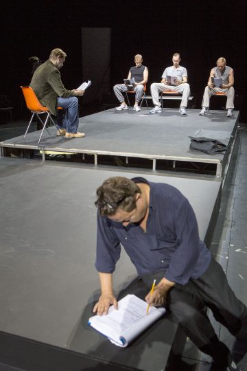 Mies polvistuu lattialla näyttämön edessä ja kirjoittaa käsikirjoitukseen. Neljä miestä istuu tuoleilla näyttämöllä, kukin keskittyneenä lukemaan tai kirjoittamaan omaan käsikirjoitukseensa. Tausta on tumma.