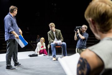 Ryhmä ihmisiä näyttämöllä harjoittelee kohtausta; yksi seisoo pitelemässä käsikirjoituksia, toinen istuu papereiden kanssa, nainen istuu lattialla katsomassa, ja kameramies kuvaa, kun taas etualalla on toinen henkilö, jolla on tatuointi.