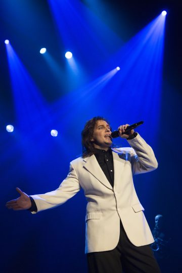 Valkoiseen bleiseriin ja mustaan paitaan pukeutunut laulaja esiintyy lavalla dramaattisessa sinisessä valaistuksessa, pitelee mikrofonia ja elehtii ojennetulla kädellä.