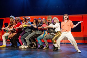 Ryhmä värikkäisiin, 1980-luvun tyylisiin pukuihin pukeutuneita esiintyjiä poseeraa yhdessä lavalla, seisoo rivissä kädet ristissä, ja taustalla on kirkas, punaisella valaistu, metrojunaa muistuttava tausta.