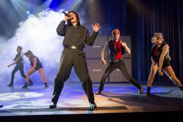 Mustiin vaatteisiin pukeutunut laulaja esiintyy intohimoisesti lavalla mikrofoni kädessään, ja häntä reunustaa neljä energistä tanssijaa dynaamisissa asennoissa, taustalla värikkäät lavavalot ja savuefektit.