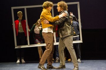 Kaksi ihmistä seisoo vastakkain ja pitelee toisiaan syleillen intensiivisessä hetkessä lavalla. Heidän takanaan kolme muuta pitää suurta suorakulmaista kehystä ja reagoi dramaattisesti. Kaikki ovat pukeutuneet värikkäisiin, retrotyylisiin vaatteisiin.