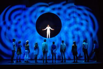 Valkoiseen pukeutunut henkilö seisoo näyttämön yläpuolella olevassa pyöreässä leikkauksessa kädet ojennettuina, kun taas ryhmä ihmisiä kohtaa hänet alapuolella. Taustalle heijastetaan sinisiä pyöriviä valoja, jotka luovat dramaattisen tunnelman.