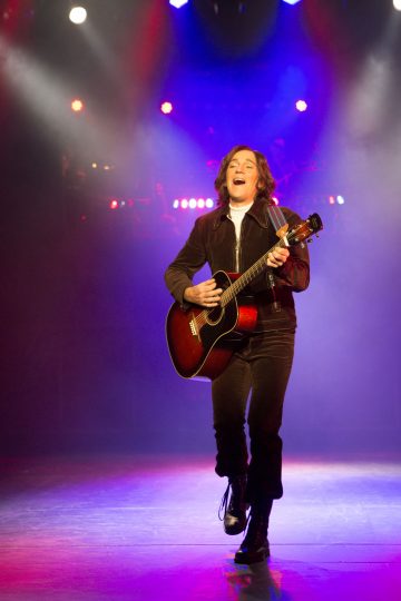 Henkilö, jolla on olkapään mittaiset hiukset, tummat vaatteet ja saappaat, soittaa iloisesti akustista kitaraa lavalla värikkäiden valojen alla.