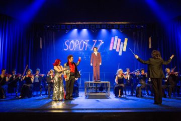 Lavaesityksessä on orkesteri, kapellimestari ja useita solisteja värikkäissä puvuissa. Yksi henkilö seisoo korotettuna näyttämön keskellä kirkkaan SOPOT 77 -kyltin alla, ja siniset valot valaisevat näyttämöä.