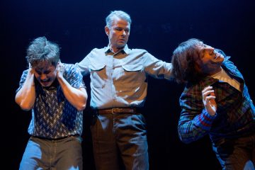 Lavalla esiintyy kolme miestä; keskellä oleva mies seisoo kahden muun välissä ja pitää heitä erillään toisistaan. Vasemmalla oleva mies peittää korvansa, kun taas oikealla oleva mies kumartuu poispäin, ja molemmat vaikuttavat ahdistuneilta dramaattisessa valaistuksessa.