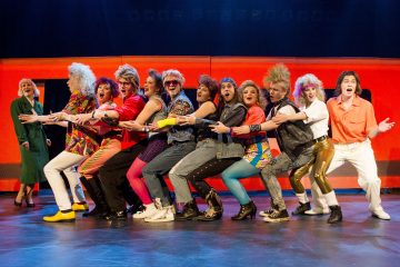 Ryhmä 1980-luvun värikkäisiin pukuihin pukeutuneita esiintyjiä seisoo lavalla riviin sidottuna, hymyillen ja kesken laulun. Kirkkaat lavavalot ja elävä oranssi tausta lisäävät energiaa vilkkaaseen kohtaukseen.