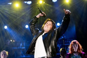 Mustaan nahkatakkiin pukeutunut henkilö laulaa lavalla innokkaasti mikrofoni kädessä ja toinen käsi kohotettuna, kirkkaat lavalamput loistavat hänen takanaan ja taustalla näkyy muita esiintyjiä.