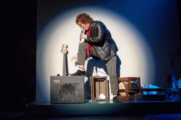 Nahkatakkiin ja raidallisiin housuihin pukeutunut esiintyjä seisoo toinen jalka Vox-vahvistimen päällä ja pitää mikrofonia lavalla valonheittimen alla. Lähistöllä on bassokitara, laatikko ja muuta lavan rekvisiittaa.