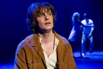 Nuori mies, jolla on ruskeat aaltoilevat hiukset ja ruskea takki, katsoo ylöspäin sinisessä näyttämövalaistuksessa, kun taustalla seisoo kaksi epäselvää hahmoa yhdessä.