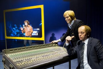 Kaksi pukumiestä, joista toinen seisoo ja toinen istuu, käyttää suurta äänimiksauskonsolia lavalla. Taustalla esiintyjä laulaa mikrofoniin keltaisella rajattujen kehysten sisällä. Kohtaus on kirkkaasti valaistu.