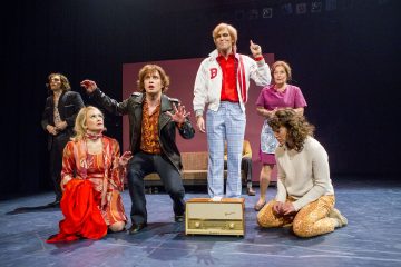 Lavalla on kuusi näyttelijää värikkäissä retropuvuissa esittämässä dramaattista kohtausta; yksi seisoo ylöspäin osoittavan radion päällä, kun taas muut polvistuvat, istuvat tai elehtivät ilmeikkäästi, ja taustalla on yksinkertainen lavastus.