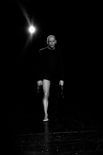 Tummapukuinen kalju henkilö kävelee lavalle paljain jaloin, kengät jalassa ja toinen jalka verkkosukkahousuissa. Kirkas valo loistaa takaa ja luo dramaattisen kontrastin.