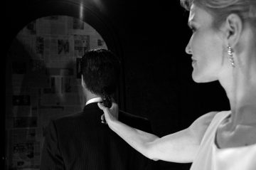 Valkoiseen mekkoon pukeutunut nainen tähtää aseella pukumiestä, joka katsoo sanomalehden peittämään peiliin ja katsoo hänestä poispäin. Kohtaus on mustavalkoinen.