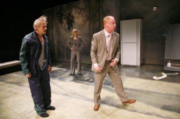 Lavalla esiintyy kolme näyttelijää. Yhdellä miehellä on tumma haalari ja hän seisoo paljain jaloin, toinen beigeen pukuun pukeutunut mies elehtii vihaisesti, ja harmaaseen pukuun pukeutunut nainen seisoo heidän takanaan. Lavasteena on jääkaappi ja maalattu tausta.