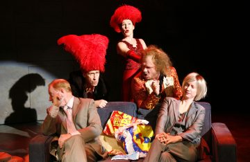 Neljä näyttelijää istuu sohvalla, kaksi harmaissa puvuissa ja tylsistynein ilmein, ja kaksi heidän takanaan, joilla on räikeät punaiset puvut ja höyhenpeitteiset hatut ja jotka reagoivat dramaattisesti. Sohvalle on ripoteltu eväspusseja.