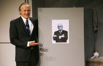 Pukumies seisoo harmaan oven vieressä, hymyilee ja osoittaa hymyillen kohti oveen kiinnitettyä mustavalkoista valokuvaa toisesta miehestä, jolla on kädet ristissä.