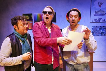 Kolme miestä seisoo sisätiloissa; keskellä olevalla miehellä on aurinkolasit ja punainen paita, hänellä on paperi kädessään ja hän laulaa. Vasemmalla olevalla miehellä on huivi ja liivi, kun taas oikealla olevalla miehellä on huppari ja värikäs pipo. Heidän takanaan roikkuu taidejulisteita.