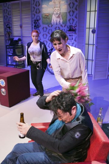 Nainen lyö vihaisesti istuvaa miestä kukkakimpulla, kun tämä pitelee olutpulloa. Toinen nainen seisoo taustalla ja katselee kohtausta. Ympäristö näyttää olevan värikäs, omituinen olohuone, jossa on eläinaiheinen sisustus.