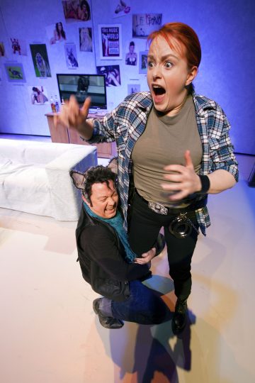 Yllättynyt punatukkainen ja ruutupaitainen nainen seisoo silmät suurina, kun kissankorvainen ja siniseen huiviin pukeutunut mies halajaa hänen jalkojaan kirkkaasti valaistussa, olohuoneen näköisessä lavasteessa.