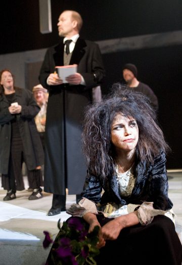 Nainen, jolla on epäsiisti tukka ja ahdistunut ilme, istuu lattialla rähjäisissä vaatteissaan, kädessään kuihtuneita kukkia, ja taustalla seisoo useita ihmisiä, muun muassa tummiin takkeihin pukeutunut mies, jolla on kädessään muistilappu.