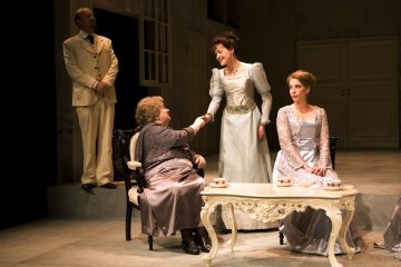 Neljä näyttelijää esiintyy lavalla aikakauden puvuissa; kaksi naista istuu koristeellisen pöydän ääressä, kun taas yksi seisoo ja kättelee istuvaa naista. Kermanväriseen pukuun pukeutunut mies seisoo heidän takanaan ja katselee. Lavastus muistuttaa eleganttia, vintage-huonetta.