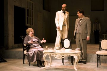 Lavalla esiintyy kolme näyttelijää: vanhempi nainen istuu tuolilla elehtimässä valkoisen teepöydän ääressä, ja kaksi pukumiestä seisoo lähellä, toinen valkoisissa ja toinen harmaissa, hämärässä, elegantissa huoneessa.