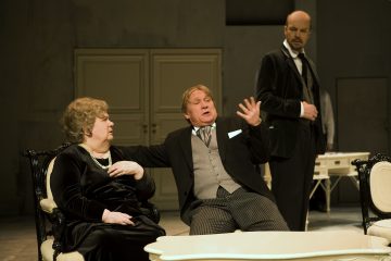 Kolme aikakauden pukuihin pukeutunutta näyttelijää esittää näyttämöllä dramaattisen kohtauksen, jossa kaksi istuu valkoisella sohvalla ja yksi seisoo heidän takanaan. Näyttämö on tyylikäs huone, jossa on klassiset huonekalut ja hillitty valaistus.