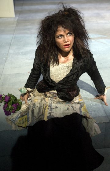 Nainen, jolla on sotkuiset hiukset ja dramaattinen meikki, istuu maassa riekaleinen asu yllään ja kädessään violetti kukkakimppu, ja katsoo ylös ahdistunut ilme.