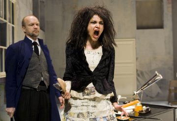 Nainen, jolla on villi tukka ja ahdistunut ilme, huutaa ja seisoo paperien peittämän työpöydän ääressä puulusikka kädessään; pukumies seisoo hänen takanaan ja katsoo.