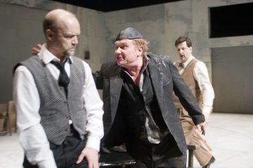 Lavalla esiintyy kolme miestä, joista toinen, jolla on lippis ja musta liivi, kohtaa vihaisesti toisen, jolla on solmio ja harmaa liivi, kun taas kolmas, ruskehtava pukuinen mies katsoo huolestuneena minimalistisessa, harmaaseinäisessä ympäristössä.