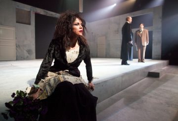 Dramaattiseen pukuun pukeutunut nainen istuu näyttämön reunalla kukkia kädessään ja näyttää ahdistuneelta. Taustalla seisoo kaksi pukuihin pukeutunutta miestä vastakkain minimalistisessa, modernissa teatterin lavasteessa.