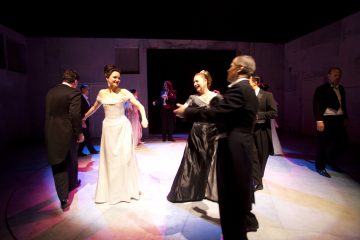 Joukko ihmisiä virallisissa iltapuvuissa tanssii yhdessä lavalla dramaattisessa valaistuksessa, ja jotkut parit pitävät toisiaan kädestä ja toiset keskustelevat taustalla.