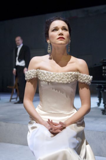 Nainen tyylikkäässä valkoisessa, helmillä koristellussa olkaimettomassa mekossa istuu kädet ristissä ja katsoo ylöspäin. Taustalla hämärästi valaistulla näyttämöllä seisoo smokkiin pukeutunut mies.