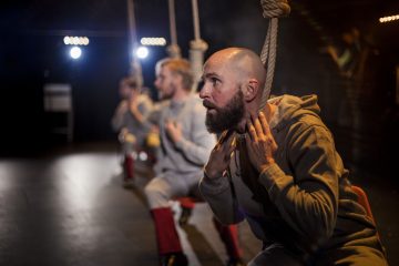 Kolme harmaisiin asuihin pukeutunutta ihmistä istuu köysistä tehdyissä keinuissa, ja etualalla on parrakas mies, joka pitää köysiä kaulansa lähellä. Taustalla loistavat näyttämön valot, jotka luovat dramaattisen tunnelman.