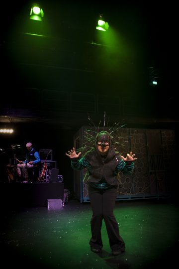 Kasvomaalattu ja piikkipäähineinen esiintyjä poseeraa dramaattisesti vihreiden näyttämövalojen alla, ja muusikko soittaa soittimia hämärässä taustalla.