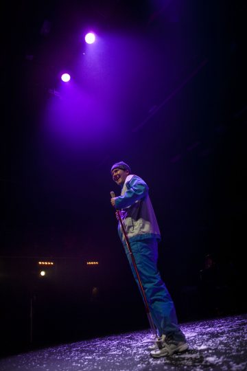 Sinisiin housuihin ja otsapantaan pukeutunut esiintyjä seisoo pimeällä lavalla violettien valonheittimien alla, keppi kädessä ja hymyillen, ja lattialle on ripoteltu valkoista konfettia.