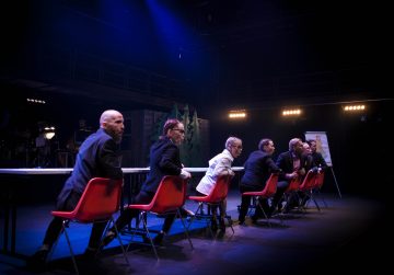 Seitsemän virallisesti pukeutunutta ihmistä istuu punaisilla tuoleilla pitkän pöydän ääressä tummalla näyttämöllä, ja kaikki katsovat sivulle vakavin ilmein. Lavavalot korostavat ryhmää, ja taustalla näkyy fläppitaulu ja maalattu lavaste.