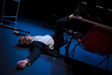 Pukuun ja silmälaseihin pukeutunut henkilö makaa selällään näyttämön lattialla toinen jalka pöydällä ja kädet ojennettuina punaisen tuolin vieressä dramaattisessa valaistuksessa.