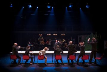 Seitsemän tummiin vaatteisiin pukeutunutta ihmistä istuu punaisilla tuoleilla pitkän pöydän ääressä lavalla, jotkut nostavat kätensä ylös, kun muusikot soittavat taustalla sinisten lavalamppujen alla.