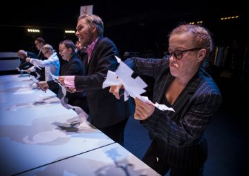 Muodollisesti pukeutunut ryhmä ihmisiä leikkaa voimakkaasti valkoisia paperinmuotoja pöydän ääressä dramaattisessa näyttämövalaistuksessa. Edessä olevalla naisella on silmälasit ja neulenauhapuku, ja hän keskittyy työhönsä.