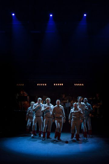 Kahdeksan univormuihin pukeutunutta näyttelijää seisoo tiiviisti yhdessä hämärästi valaistulla näyttämöllä, jota valaisee ylhäältä tuleva sininen valonheitin, ja heidän takanaan varjoissa näkyvät muusikot ja soittimet.