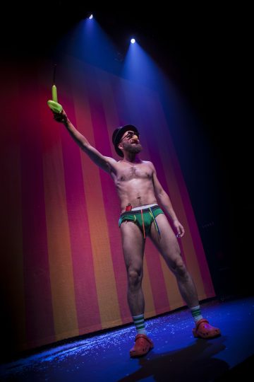 Parrakas mies värikkäissä alusvaatteissa, sukissa ja punaisissa kengissä seisoo lavalla sinisten valojen alla ja pitää vihreää rekvisiittaa ylhäällä. Hänellä on aurinkolasit ja hattu, ja hänen takanaan on dramaattinen oranssin ja vaaleanpunaisen raidallinen tausta.