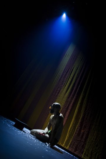 Uimalaseja käyttävä henkilö polvistuu yksinään hämärästi valaistulla näyttämöllä sinisen valonheittimen alla, taustalla on värikkäitä pystyraitoja ja lattialle on ripoteltu konfetteja.