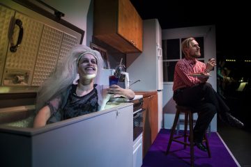 Häähuntuun pukeutunut nainen istuu hymyillen avonaisessa pakastimessa juoma kädessään, kun punaruutuiseen ruutupaitaan pukeutunut mies syö läheisellä jakkaralla kirkkaasti valaistussa keittiön lavastuksessa.