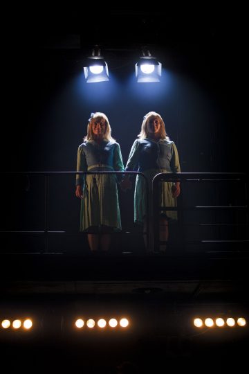 Kaksi vihreisiin mekkoihin pukeutunutta naista seisoo vierekkäin pimeällä näyttämöllä kädestä pitäen kirkkaiden valonheittimien alla, ja heidän kasvonsa ovat osittain valaistut.