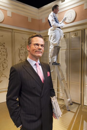 Etualalla hymyilee mies, jolla on nastaraitapuku ja vaaleanpunainen solmio, ja hänellä on kädessään taiteltuja sanomalehtiä. Taustalla toinen henkilö työskentelee tikkailla koristeellisen seinälistan parissa tyylikkäässä huoneessa.