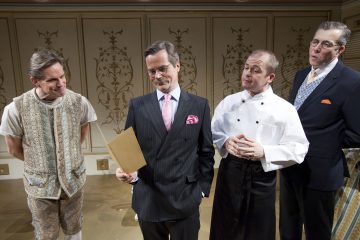 Neljä miestä seisoo sisätiloissa: yhdellä on kuviollinen liivi, yhdellä pukumies, jolla on kansio kädessään, yhdellä kokin univormu ja yhdellä pukumies, jolla on silmälasit. He näyttävät keskustelevan tai osallistuvan esitykseen. Taustalla on koristeltu tapetti.